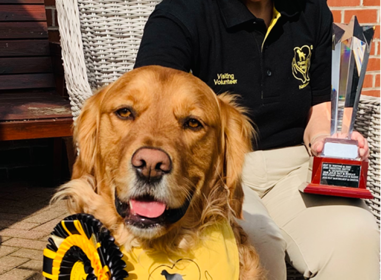 Therapy dog, Chester, with a yellow and black rosette. Owner Kay holds a trophy