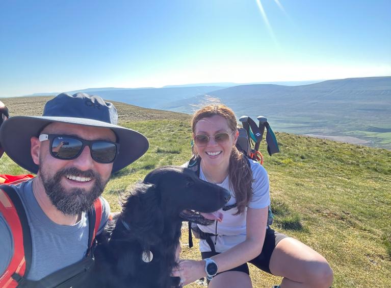 A man and woman with a black dog with a remote hilly background