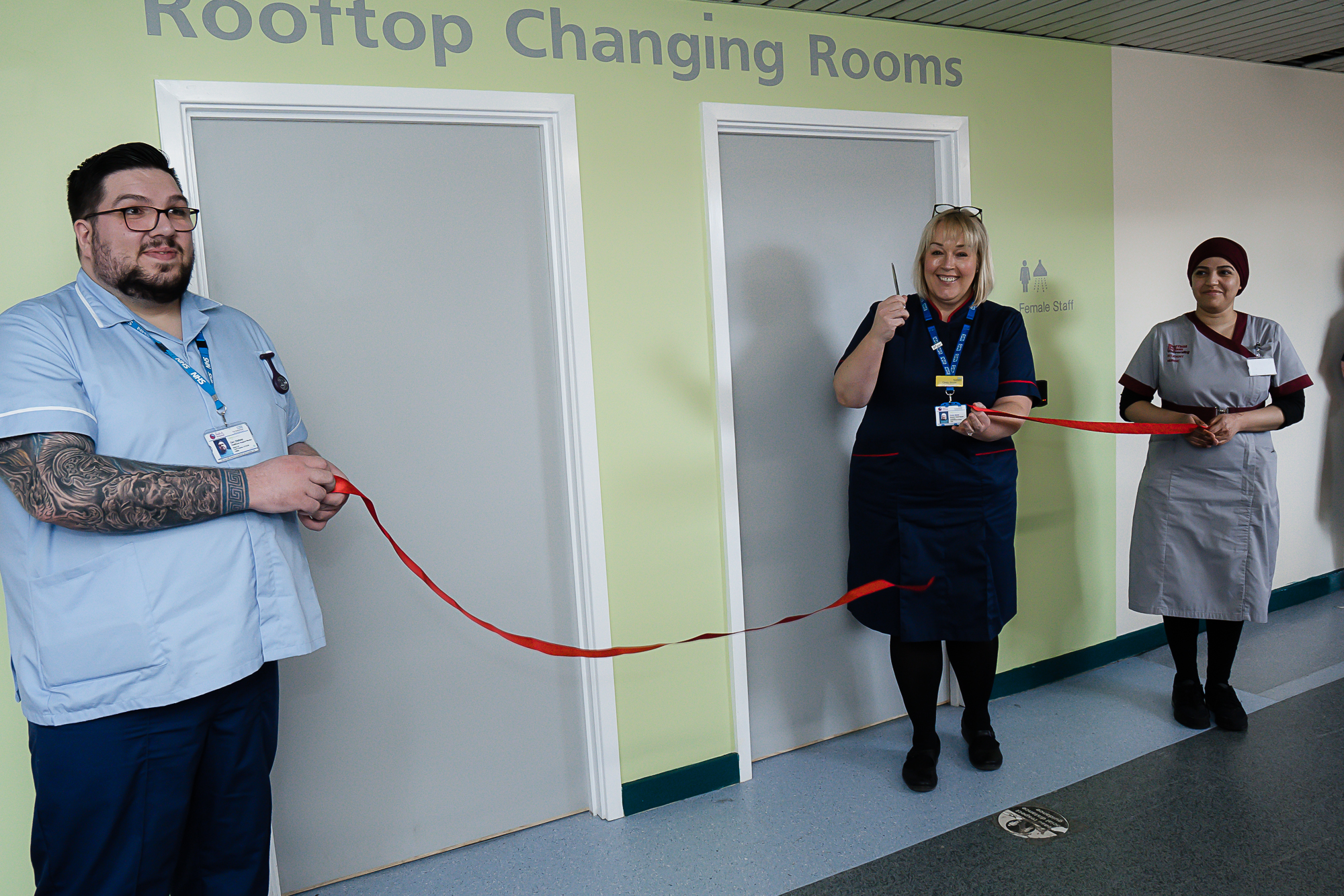 Chief Nurse, Cindy Storer, cuts a red ribbon held by a healthcare support worker and student nurse to open some new changing rooms