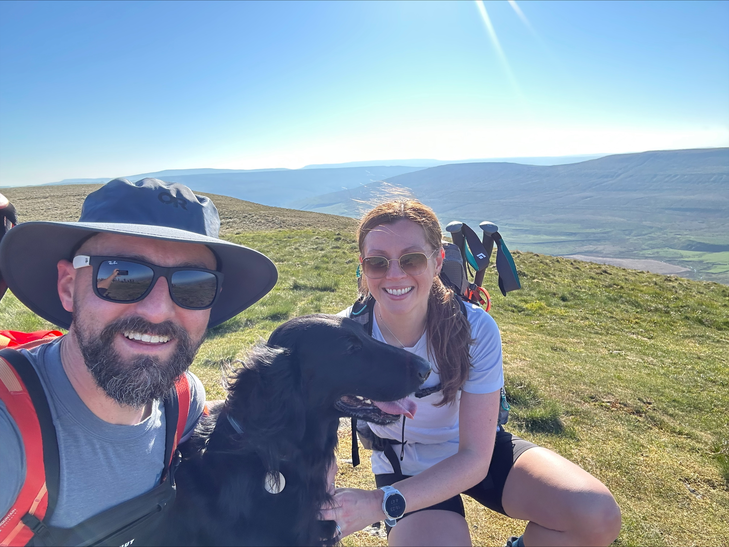 A man and woman with a black dog with a remote hilly background