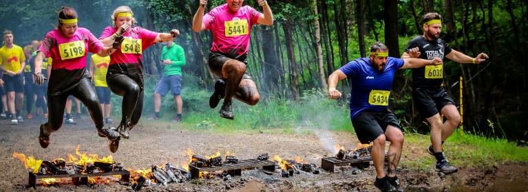 Five people, three in pink charity t-shirts, jump over fire into a muddy puddle