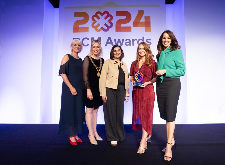 Kayty is wearing a dark orange dress and is holding a trophy. There are three people to her left and one to her right.