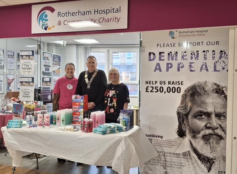 Three people are stood behind a table with toiletries in front of Rotherham Hospital and Community Charity's Hub.
