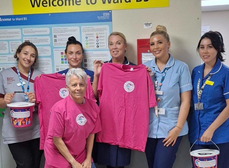 Six women are smiling for the camera. Three have pink charity-branded t-shirts and two are holding pink charity collection buckets
