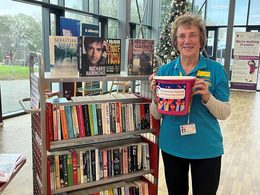Volunteer in a blue t-shirt holding a pink charity collection bucket beside shelves of books