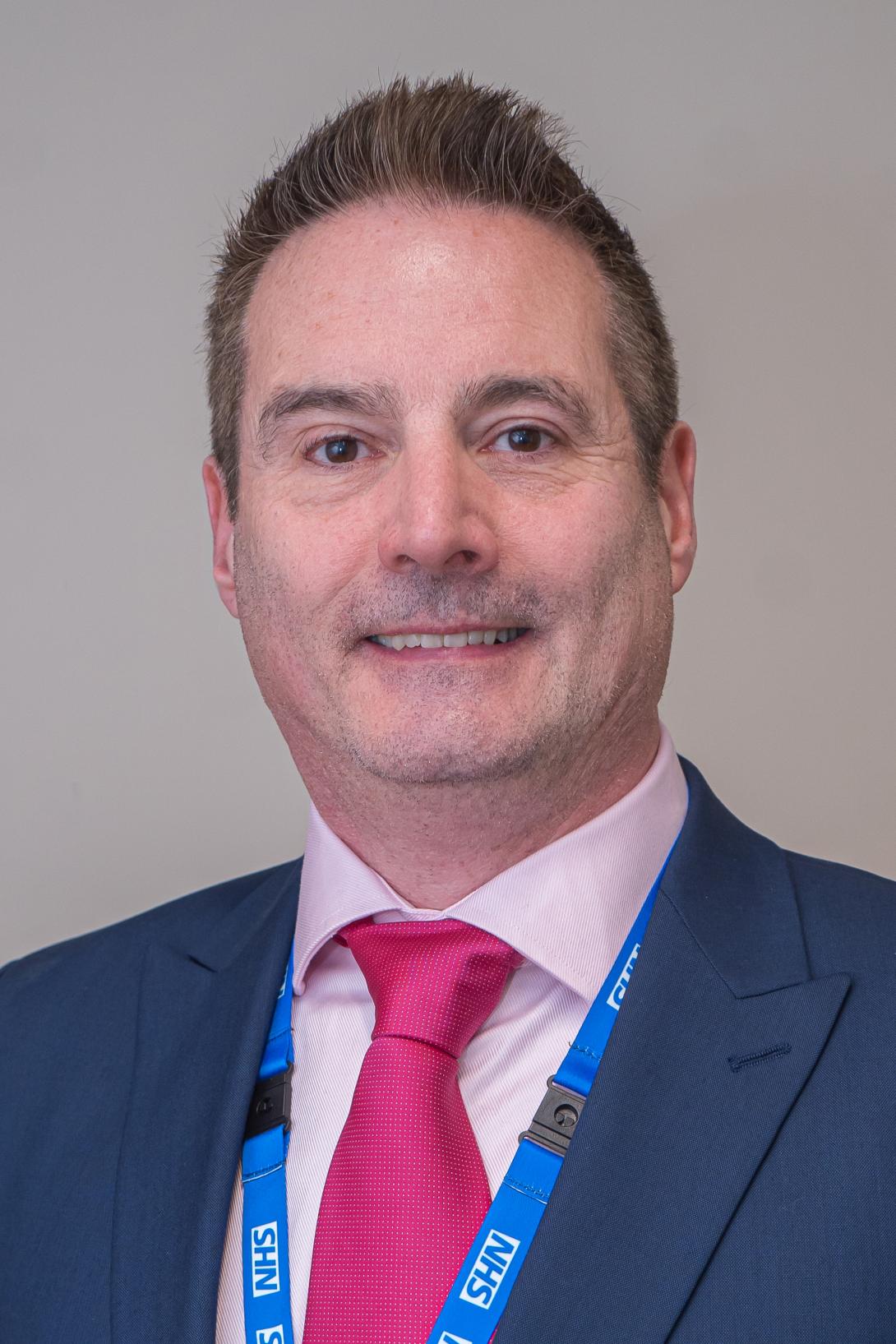 Shane is smiling at the camera. Hi is wearing a blue suit jacket with a light pink shirt and a dark pink tie.
