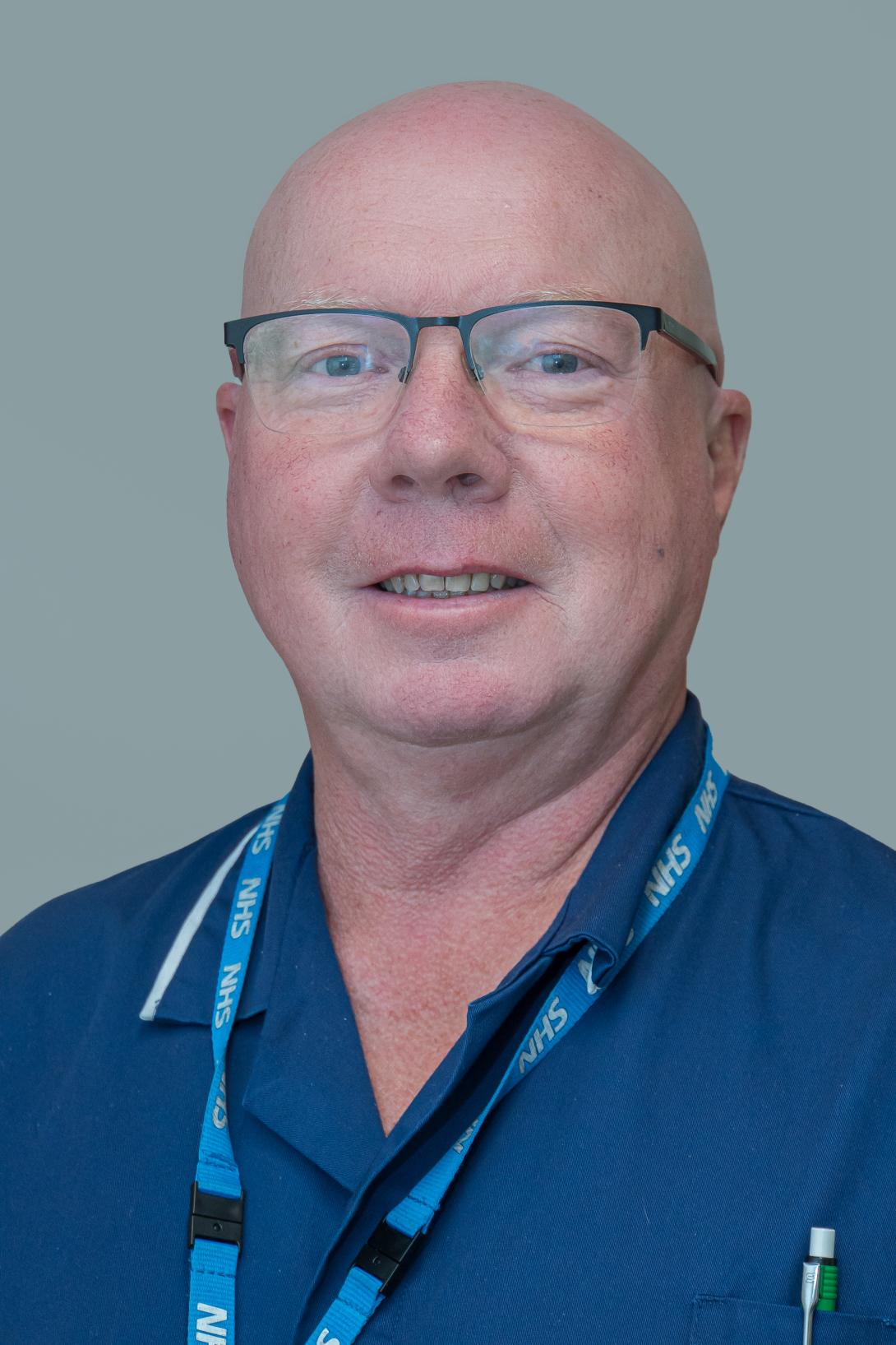 Andrew is wearing glasses and a blue nursing uniform with blue NHS lanyard